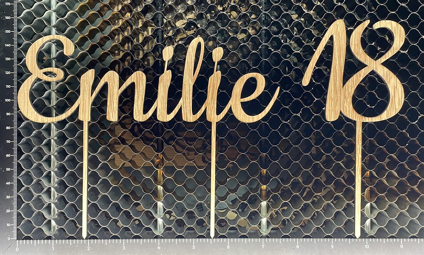 Frisch ausgeschnittener Tortenstecker mit dem Schriftzug „Emilie 18“ auf dem Wabentisch des Lasercutters in der Werkstatt in Lohnsburg